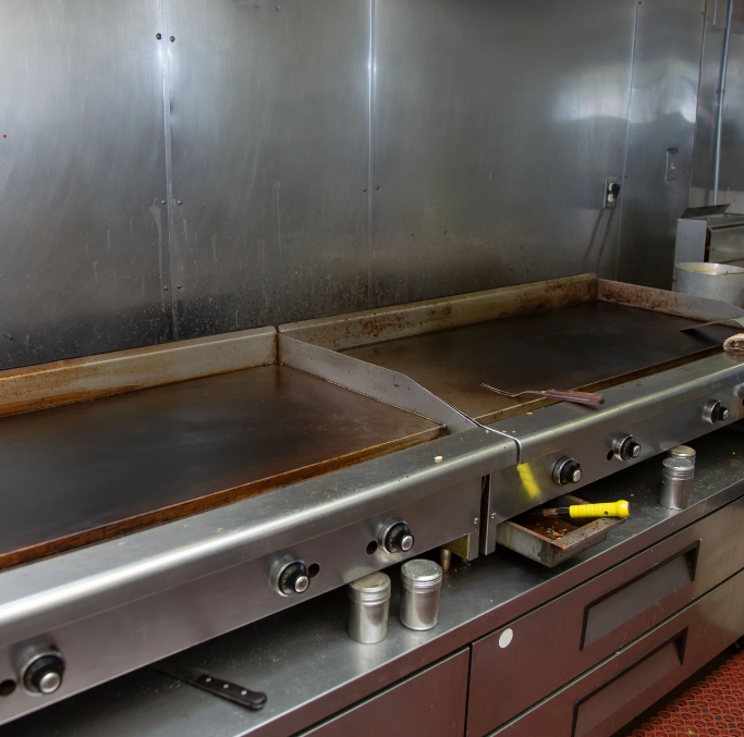Commercial kitchen flat-top griddle with two dark cooking surfaces and stainless steel backsplash