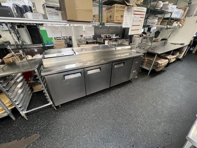 Stainless steel prep counter in a commercial kitchen with shelves, supplies, and a speckled black floor