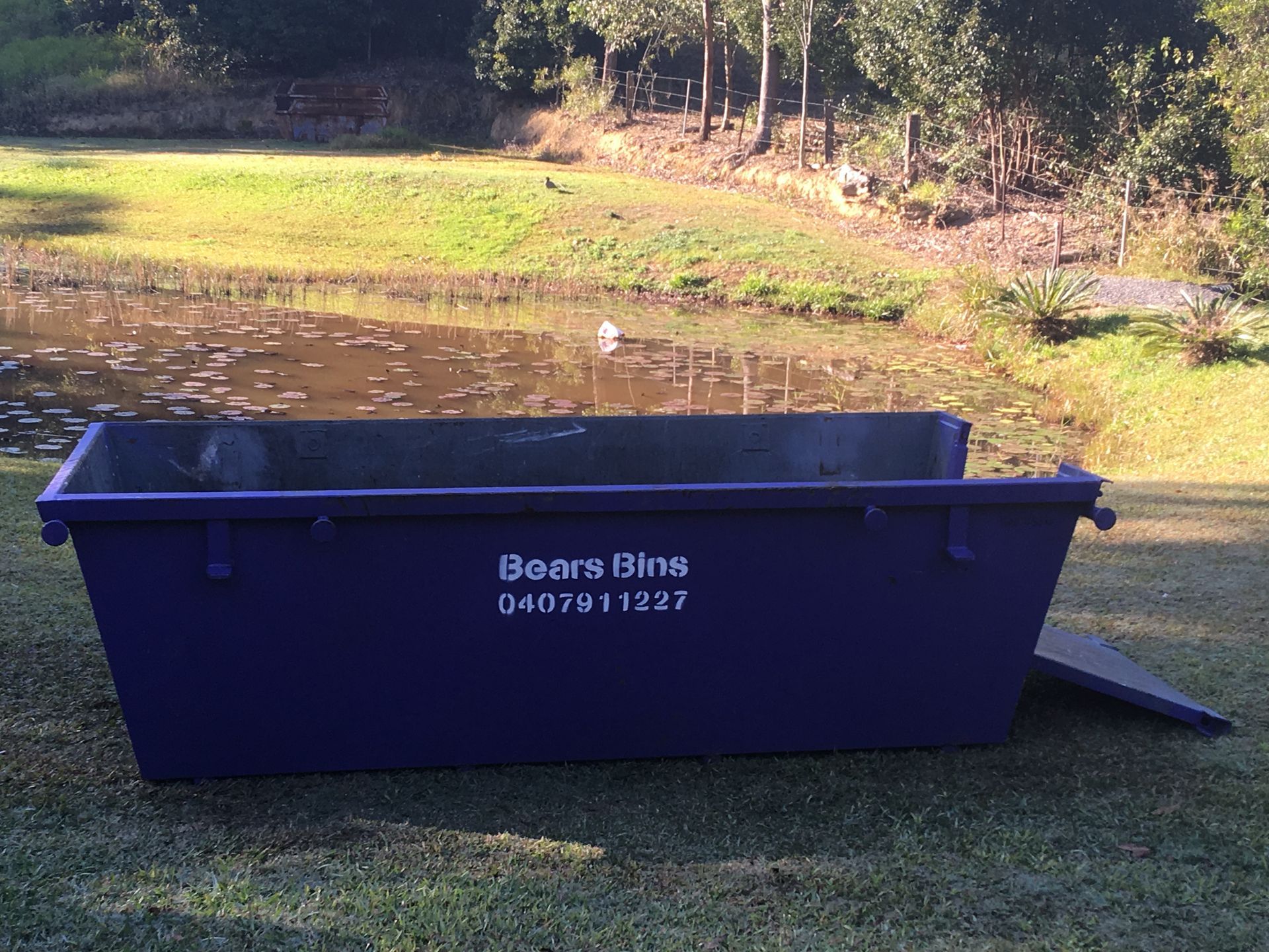Blue skip bin on grass beside a pond, with