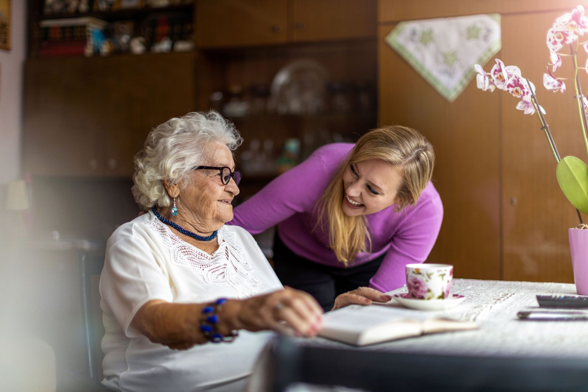 Woman leaning toward smiling elderly woman at a dining table with a cup and flowers in the foreground