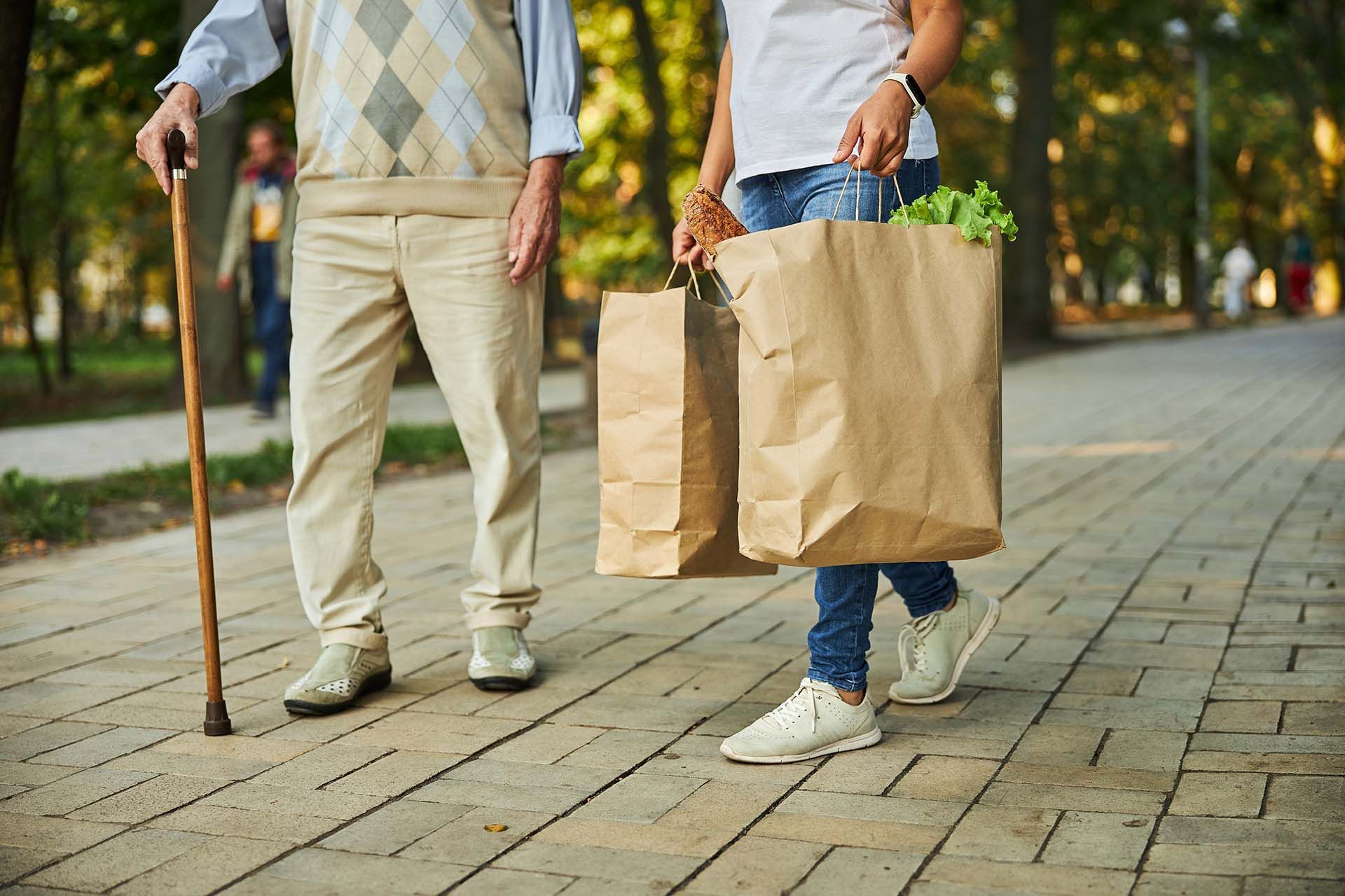 A caregiver holding paper shopping bags and walking with an elderly man using a cane outdoors.