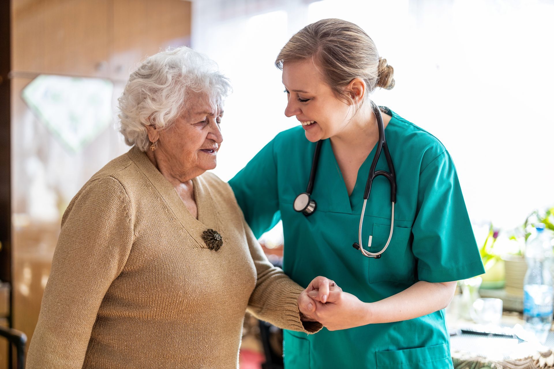 Nurse in teal scrubs gently supports an elderly woman indoors, both smiling during a care visit