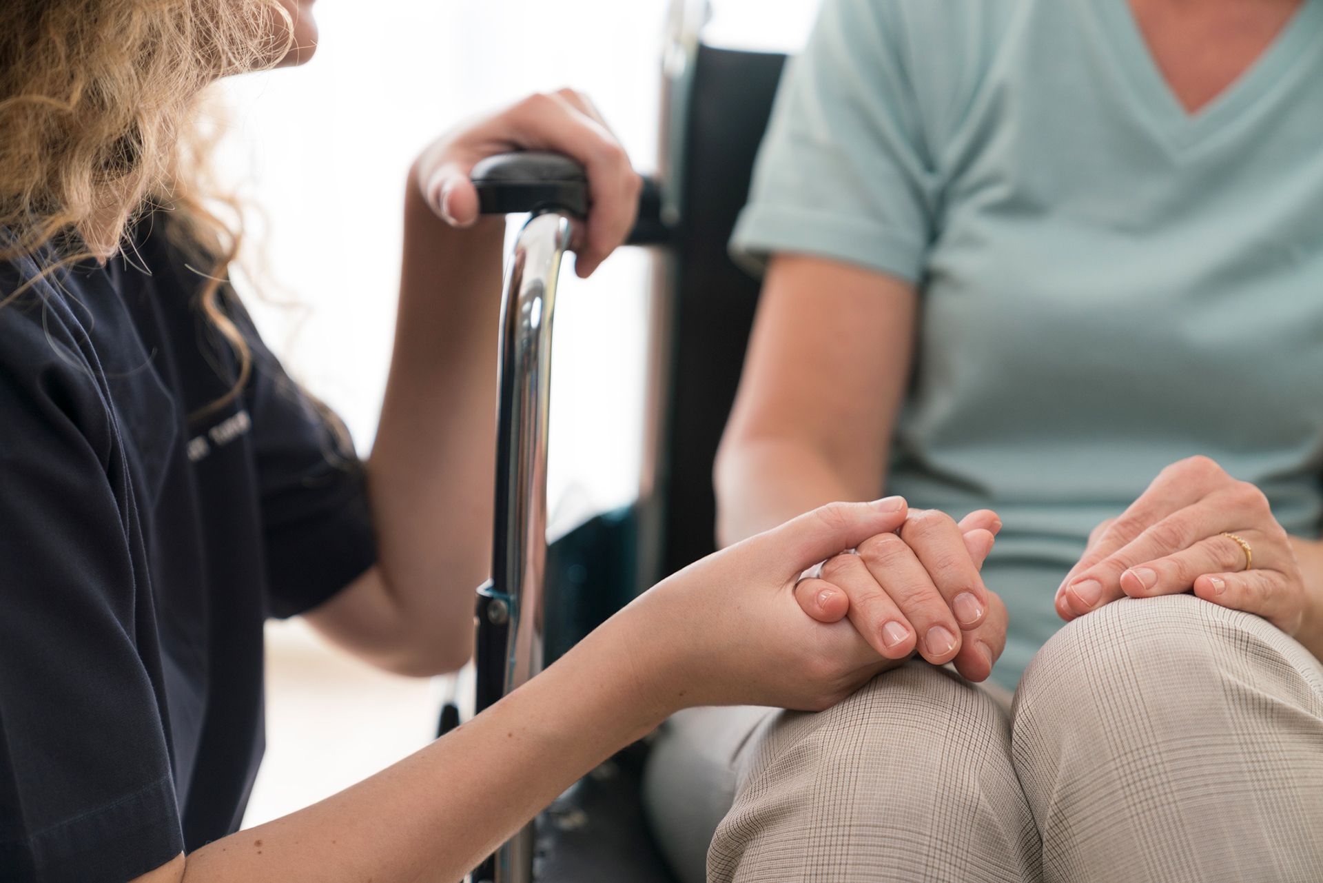 A home nurse is holding the hand of a senior woman in a wheelchair.