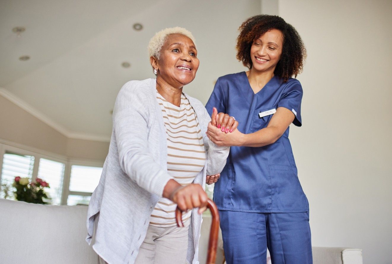 Woman Using Cane, Assisted By A Healthcare Worker. Both Are Smiling Indoors.