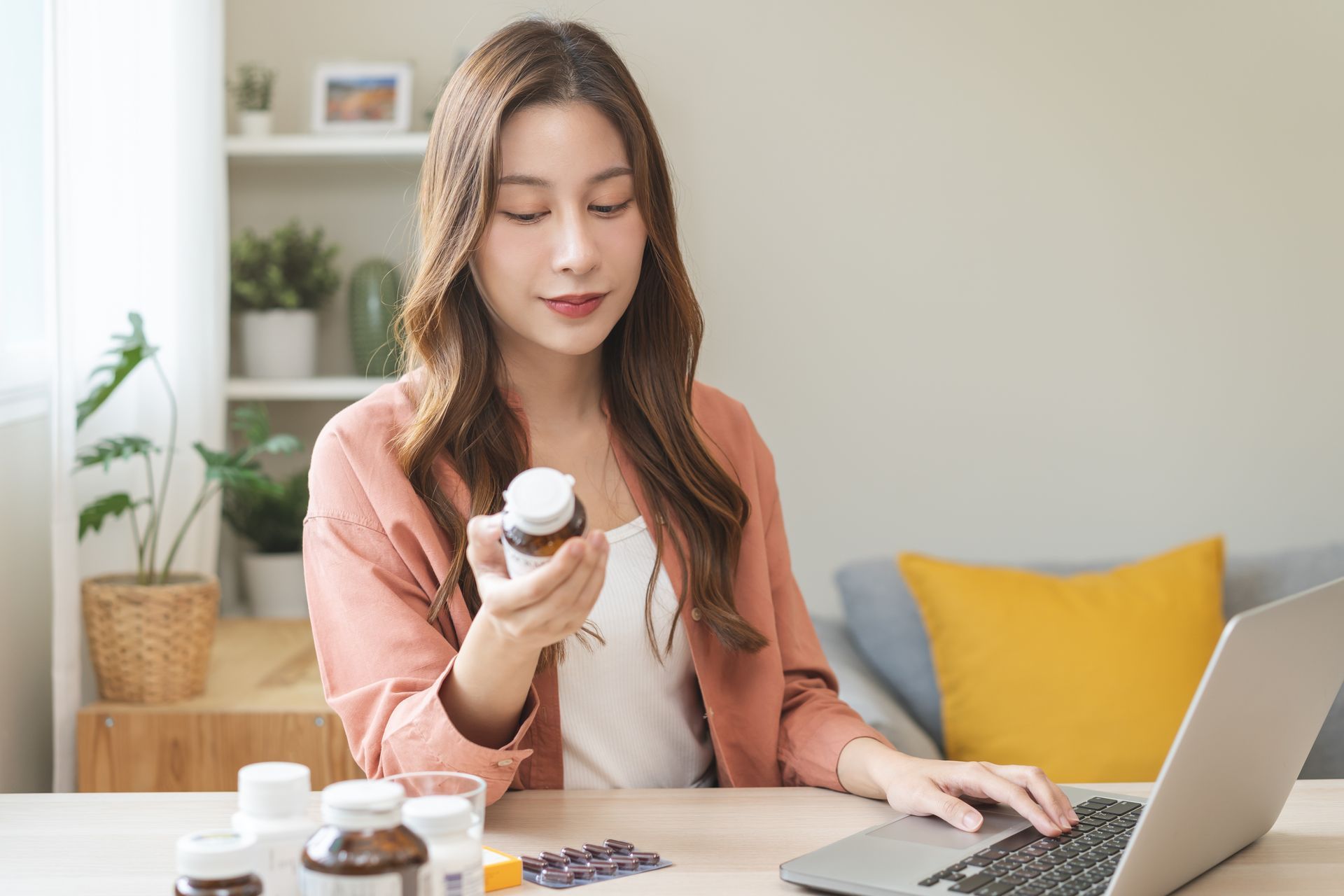 A woman is sitting at a table holding a bottle of supplements and using a laptop computer to research supplements for fertility