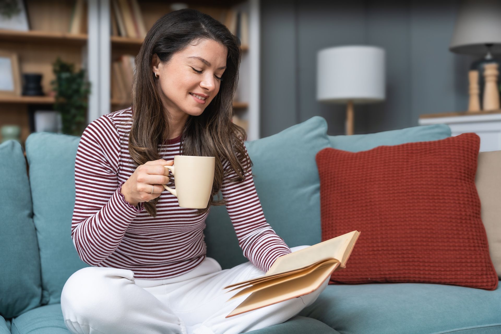Woman reading on a teal couch while holding a mug, with a red pillow and lamp in the background