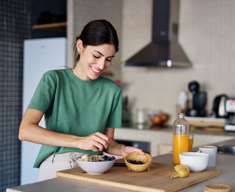 Person smiling while preparing breakfast in a kitchen, with bowls, fruit, and a glass of orange juice.