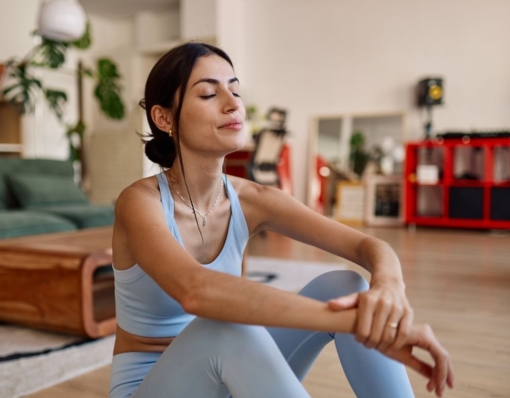 Woman in light blue activewear sitting on a rug, eyes closed, in a bright living room.