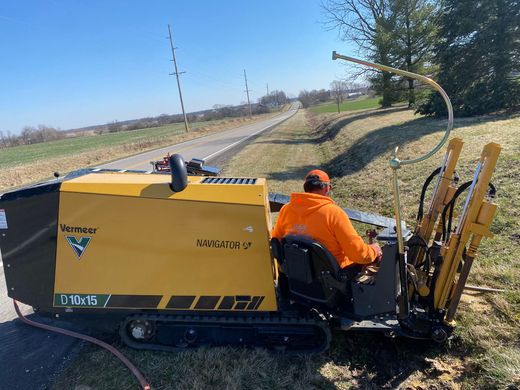 Orange utility worker riding a yellow tracked machine along a rural roadside beside a field