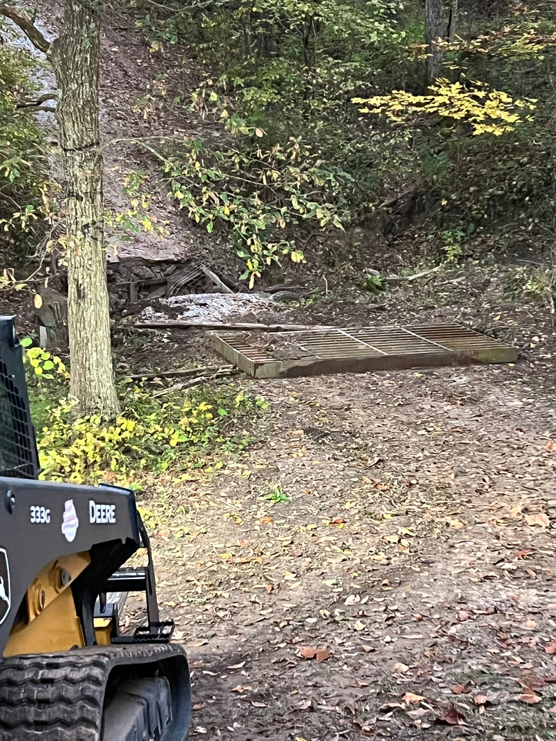 Wooded trail with a narrow stream crossing and a partially visible excavator in the foreground
