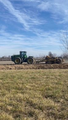 Green tractor plowing a field under a blue sky, with dry grass in the foreground