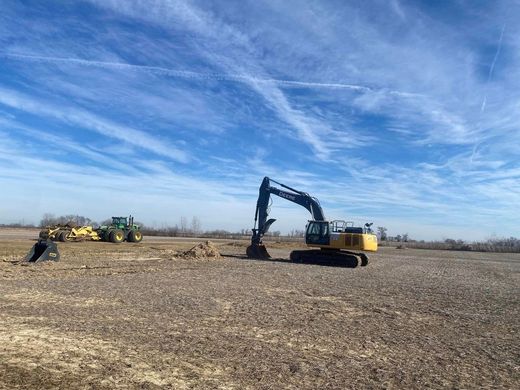 Yellow and black excavator on a dry field under a blue sky, with a tractor in the distance.