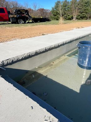 Concrete pool with clear water and a blue bucket beside it; red truck and trees in the background.