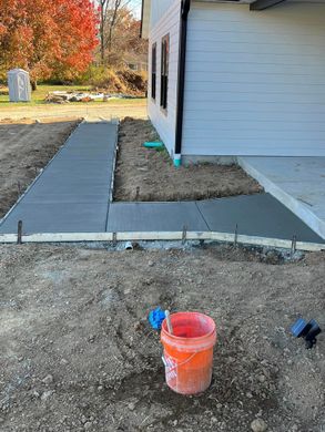 New concrete walkway and patio beside a white house, with gravel and an orange bucket in the foreground.
