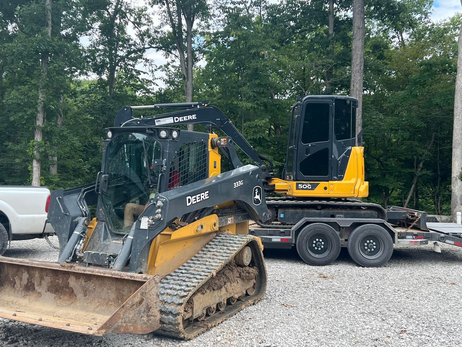 Black skid steer on trailer beside a pickup in a wooded area