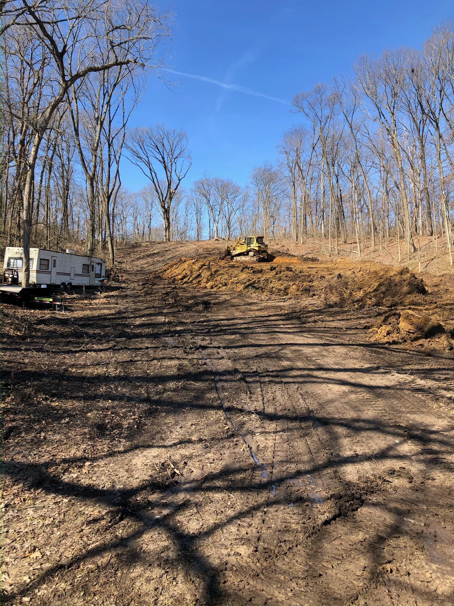 Cleared dirt road through bare trees under blue sky, with a yellow bulldozer in the distance