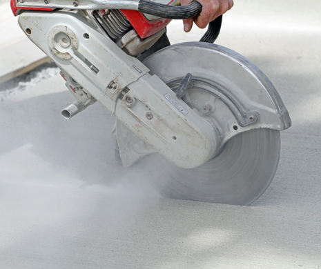 Worker using a concrete saw to cut pavement, with dust cloud rising.