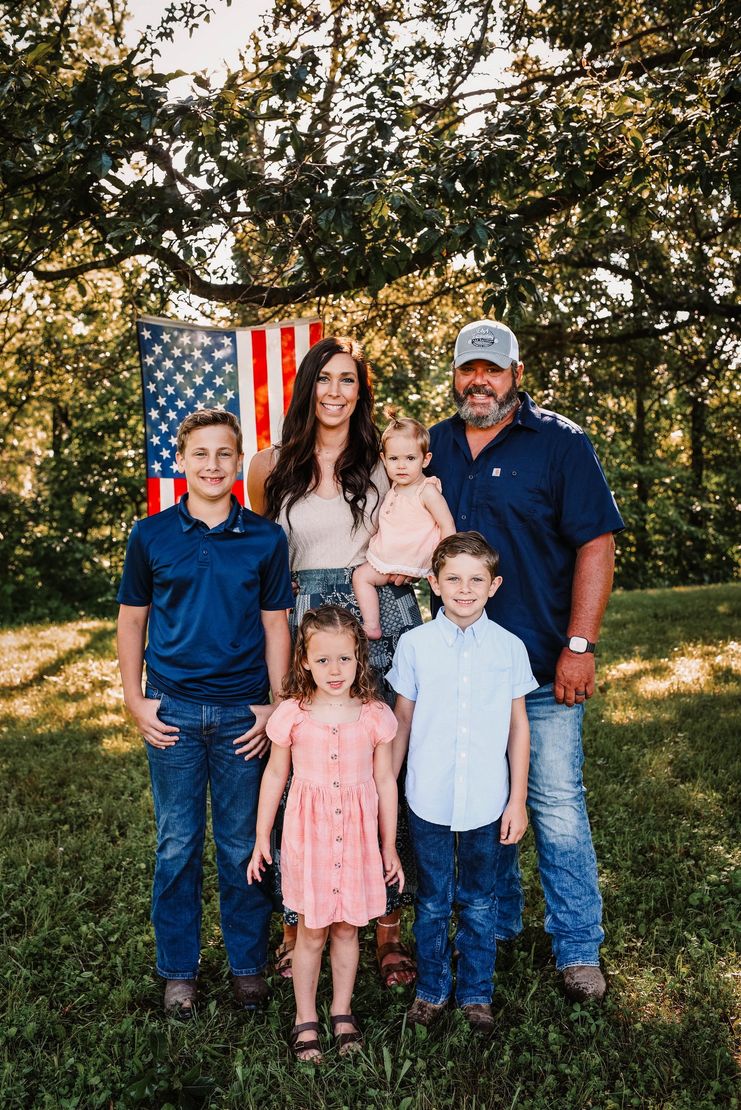 Family of five posing outdoors in front of an American flag and trees, smiling at the camera.