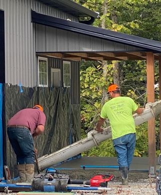 Workers pouring concrete near a house extension, using a long chute under a covered porch.
