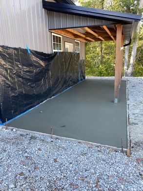 Covered concrete patio beside a building, bordered by black privacy tarp and gravel landscaping.