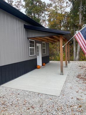 Front porch of a gray house with a pumpkin and an American flag, surrounded by gravel and autumn trees.