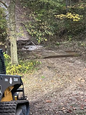 Wooded path with fallen leaves, a concrete culvert, and a partial construction loader in the foreground