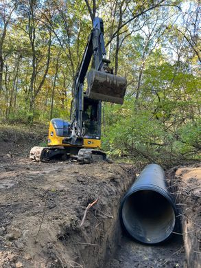 Excavator placing a large concrete culvert pipe in a wooded trench