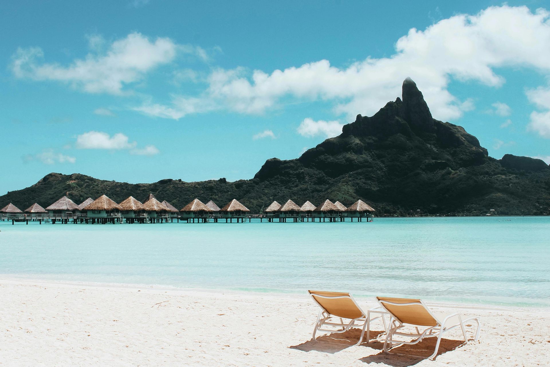 Tropical beach with turquoise water, overwater huts, rocky island, and two lounge chairs on white sand
