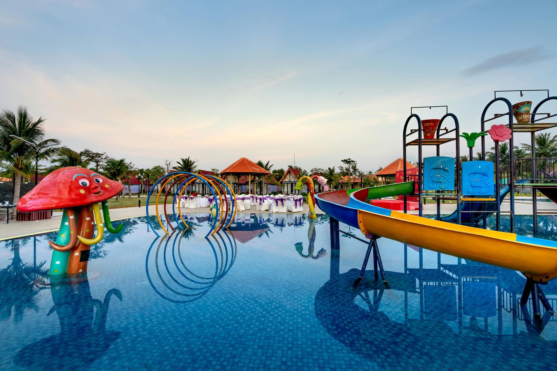 Colorful water park splash area with mushroom fountain, slides, and pools at sunset
