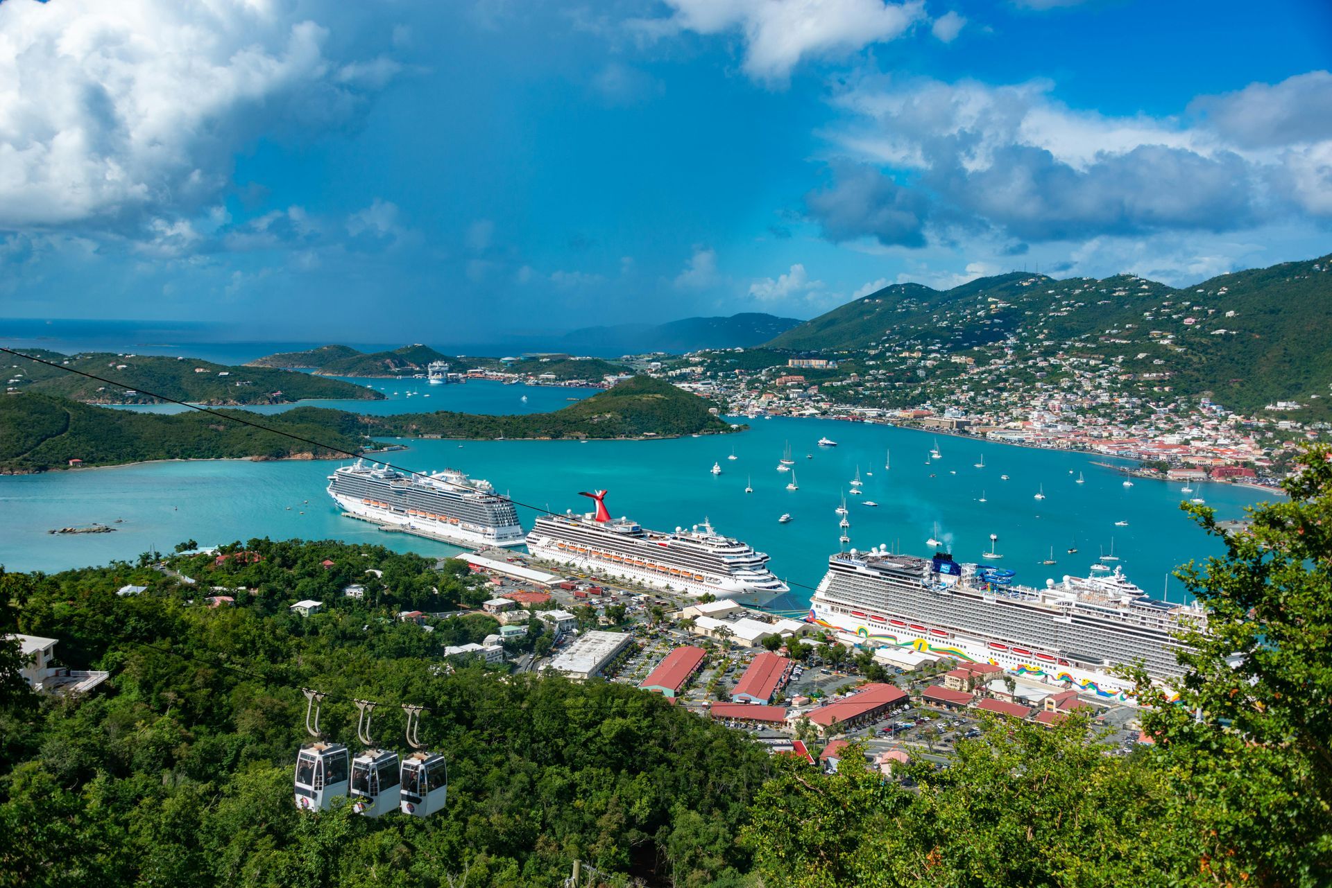 Aerial view of a turquoise bay with cruise ships, sailboats, and a coastal town under a cloudy blue sky