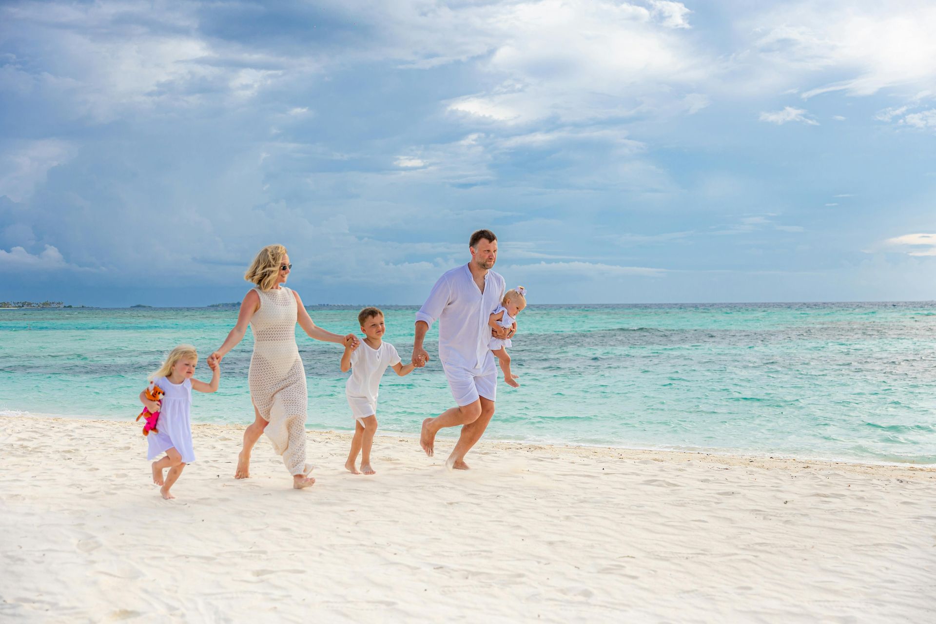 Family running hand-in-hand on a white sandy beach beside turquoise water under a cloudy sky