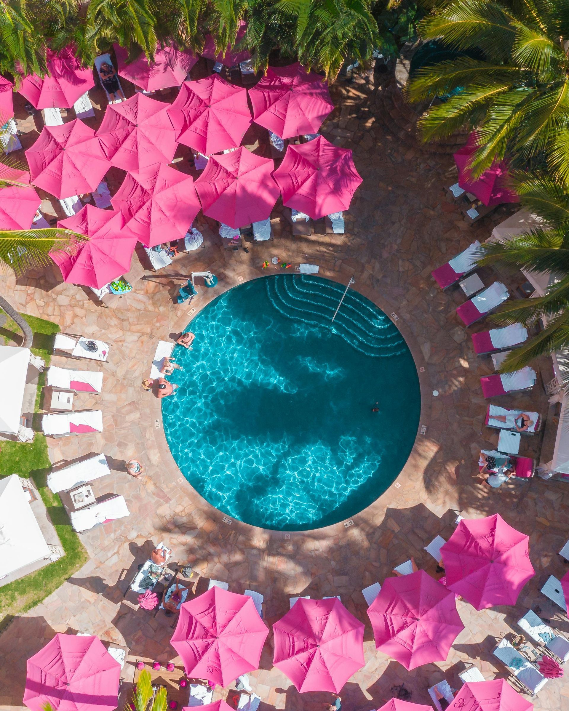 Overhead view of a circular pool surrounded by pink umbrellas and lounge chairs in a sunny resort courtyard