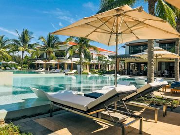 Luxury resort pool with lounge chairs, beige umbrellas, and palm trees under a bright blue sky