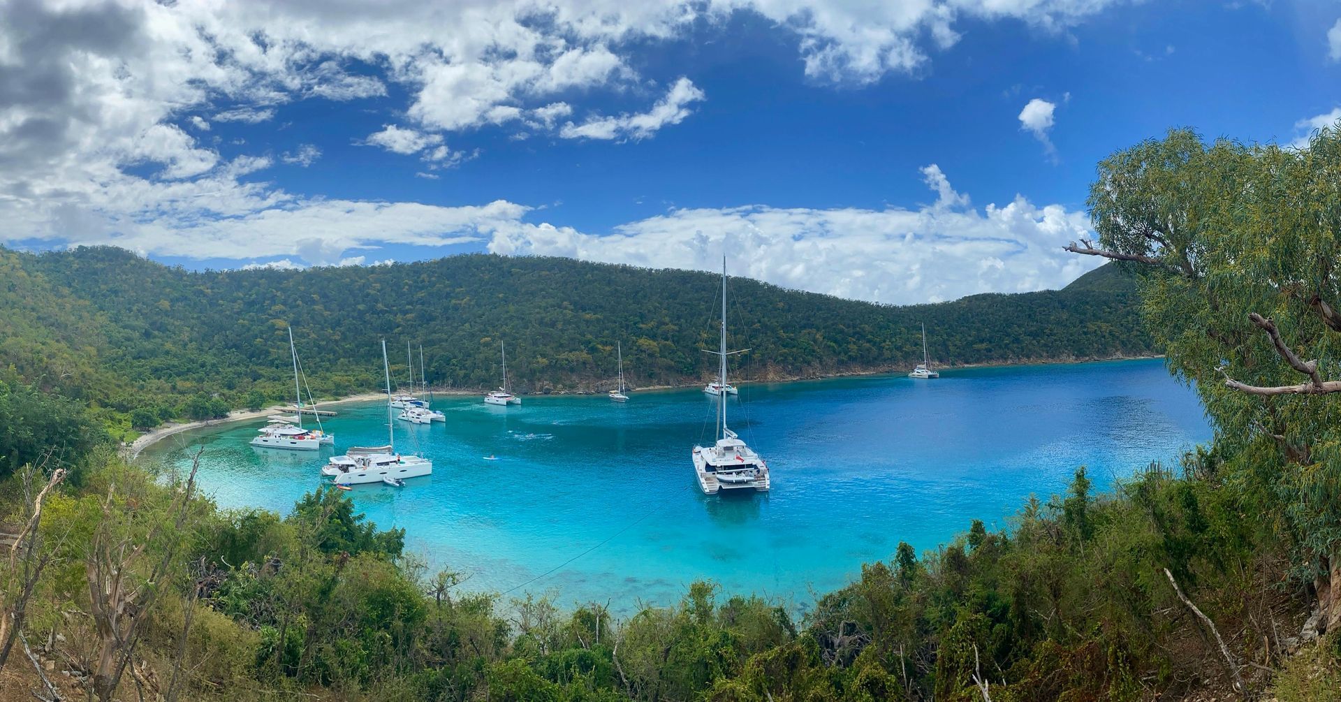 Turquoise bay with anchored sailboats, green hills, and blue sky with clouds