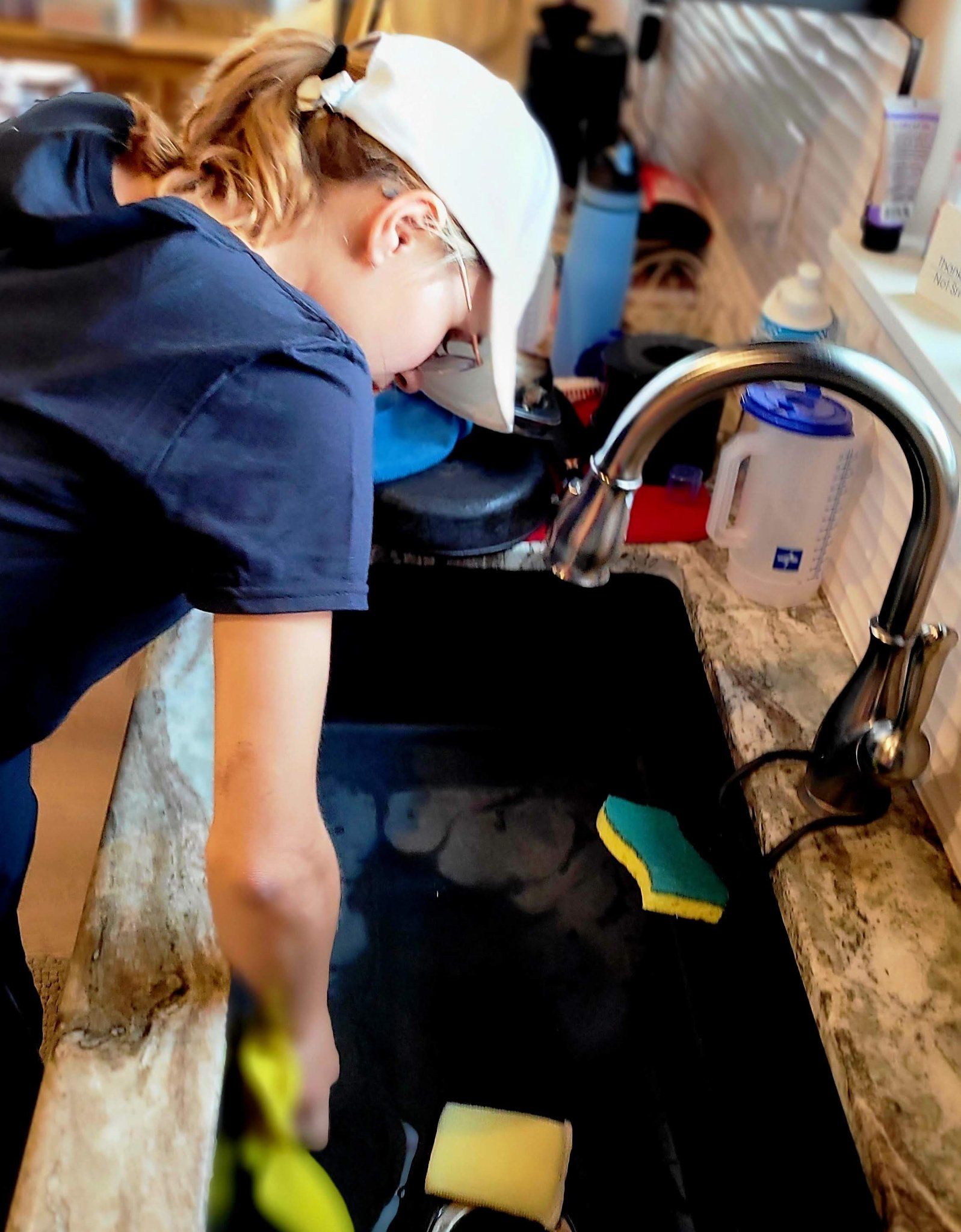 A woman is cleaning a kitchen sink with a yellow sponge.