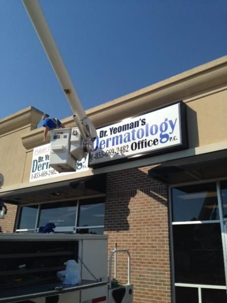 A person in a bucket truck works on a sign for Dr. Yeoman's Dermatology office mounted on a brick building exterior.
