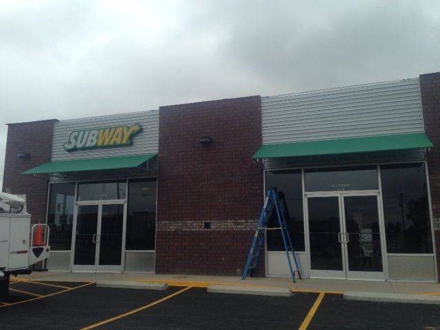 A Subway restaurant in a strip mall with a blue ladder leaning against the vacant storefront next to it.