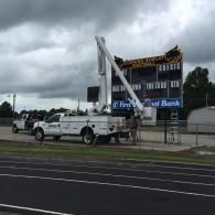 A utility truck with a raised boom bucket positioned to work on a football stadium scoreboard under a cloudy sky.