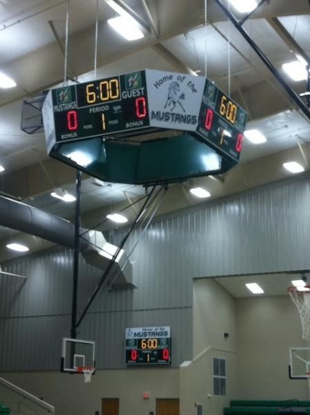 A basketball court scoreboard hanging from the ceiling showing 6:00 on the clock and 0-0 scores, with a wall unit below.