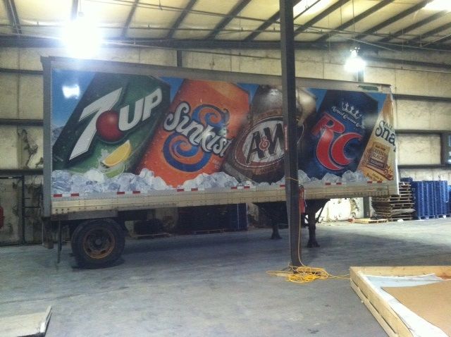 A side view of a parked semi-trailer in a warehouse, featuring large colorful graphics of 7 Up, Sunkist, A&W, and RC Cola.
