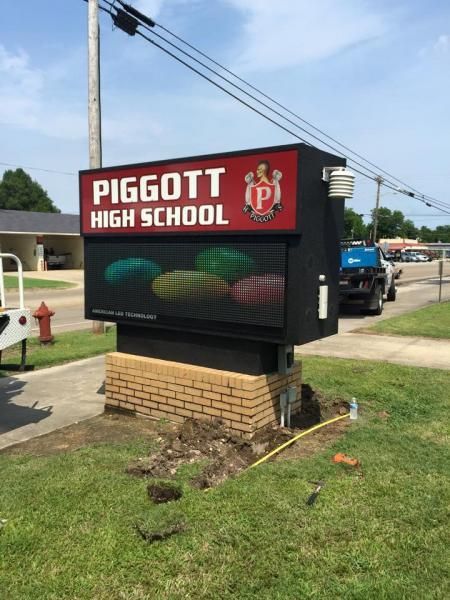 A Piggott High School outdoor electronic sign on a brick base, standing on a lawn near a road with a parked truck.