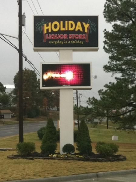 A two-tiered sign for Holiday Liquor Store, featuring a black top sign and an illuminated digital screen below.