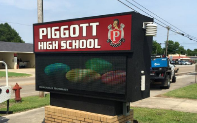 Piggott High School sign with a red panel and LED display screen on a brick base next to a road.