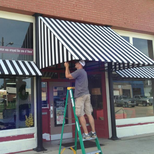 A person stands on a green stepladder installing a black-and-white striped awning over a storefront entrance.