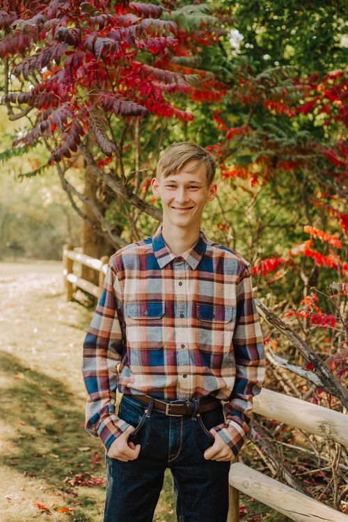 Young person in plaid shirt and jeans, smiling, standing near a wooden fence and fall foliage.