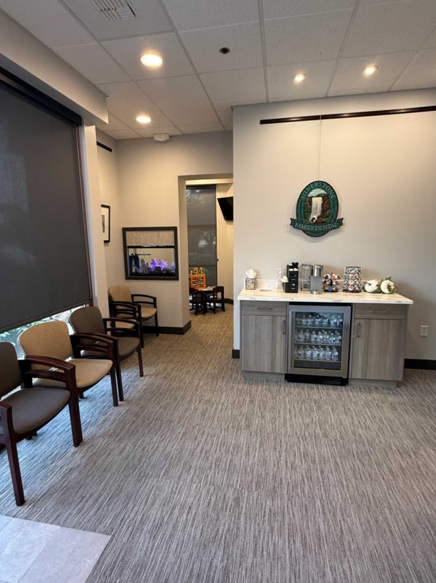Waiting area with chairs, coffee station, aquarium, and doorway. Light gray carpet and walls.