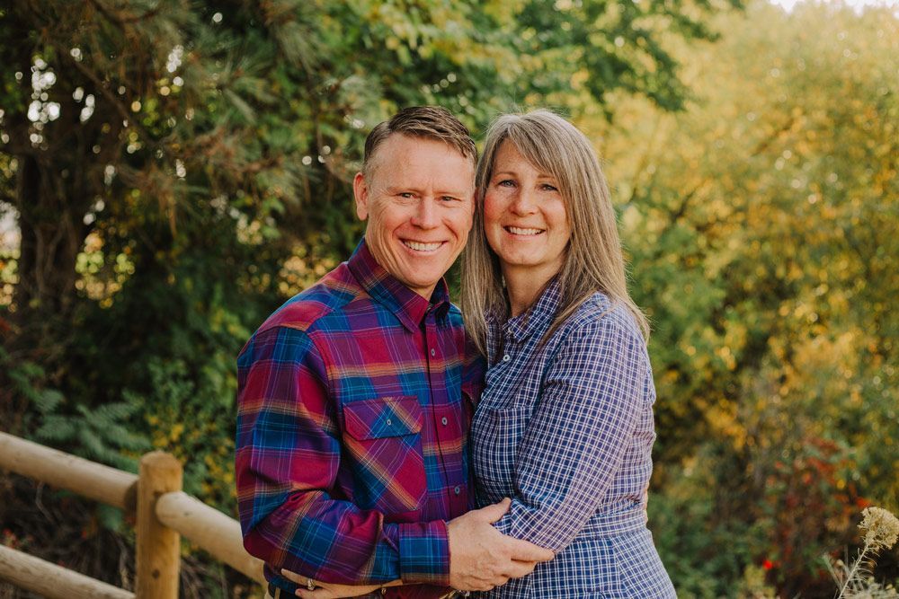 Couple smiling, embracing outdoors. Man in plaid shirt, woman in blue, both in front of foliage and fence.