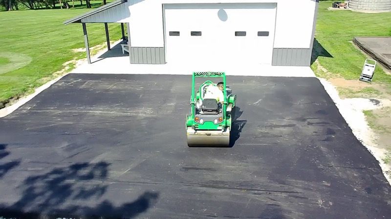 Green skid steer on a freshly paved driveway in front of a white garage.