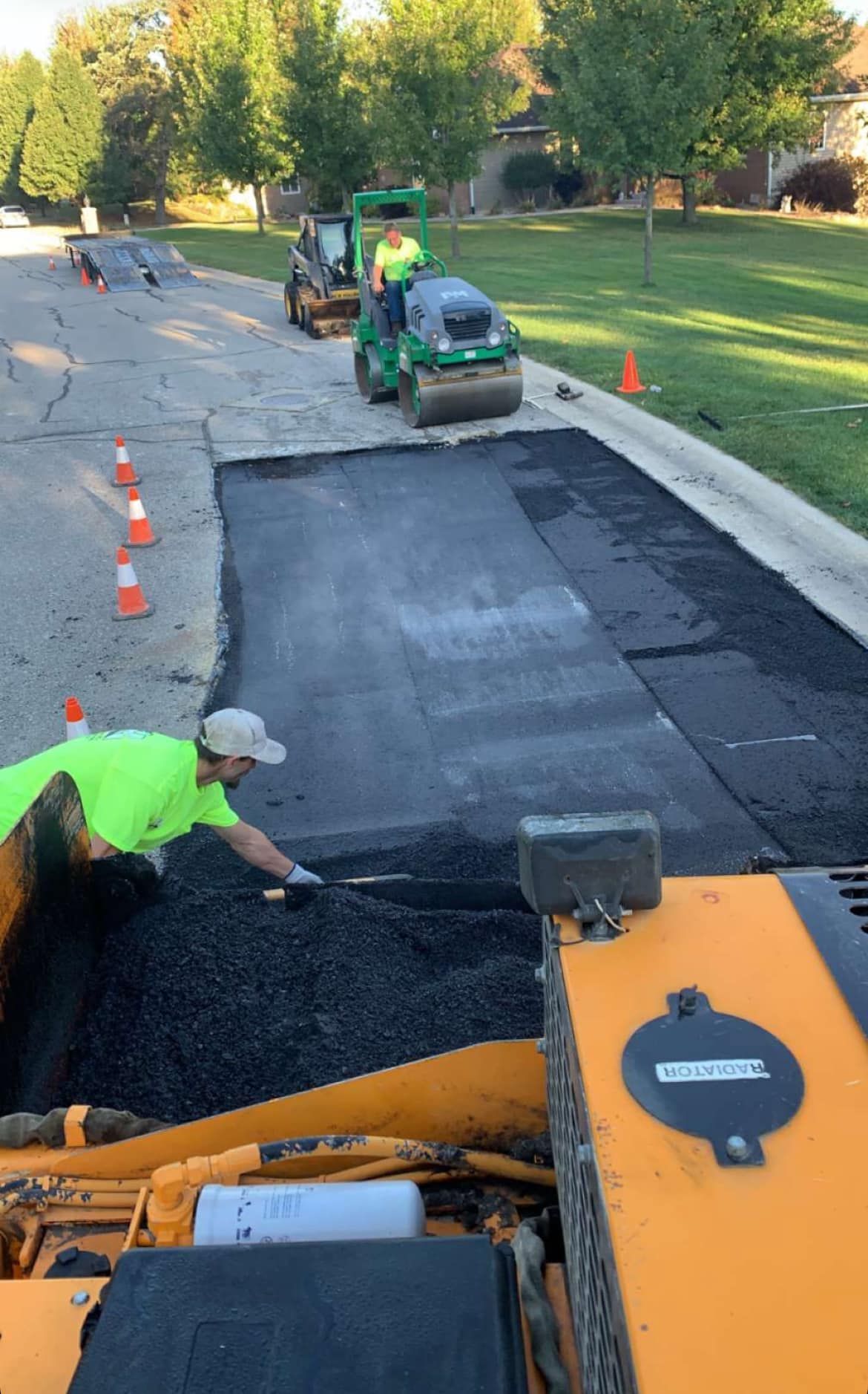 Road workers repaving a street, with a roller compacting fresh black asphalt beside orange cones.