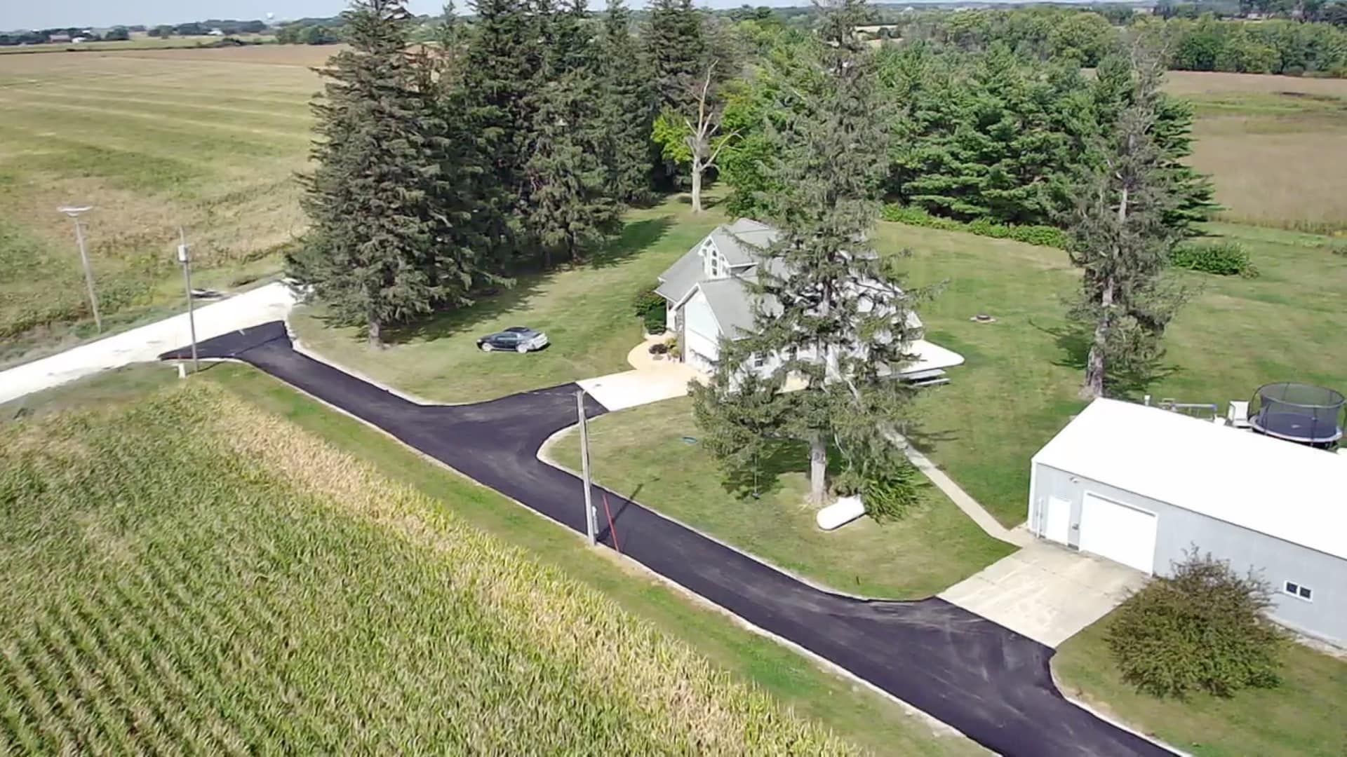 Aerial view of a farmhouse with white outbuildings, black driveway, and surrounding fields and trees.